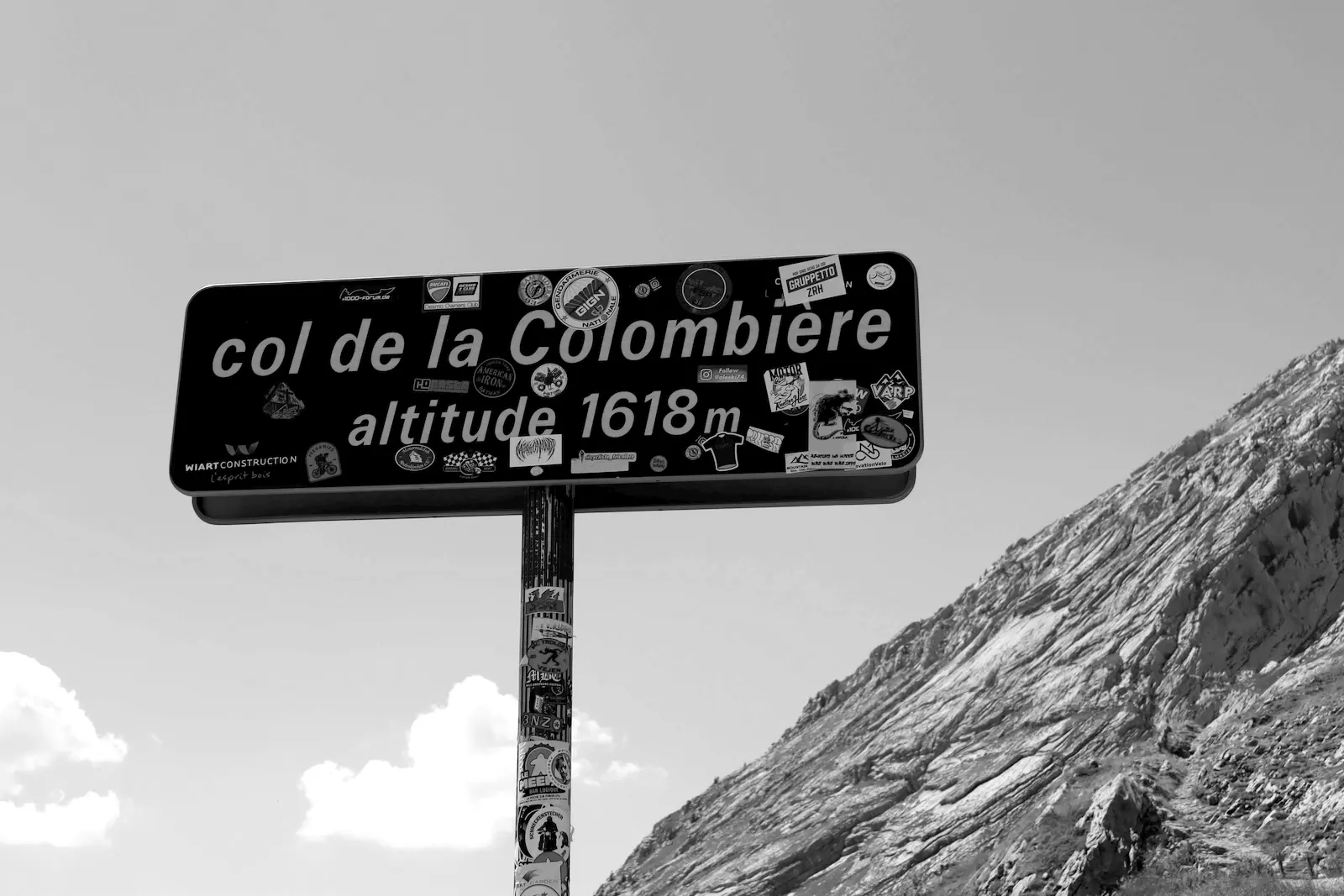 The summit sign at the Col de la Colombière, reading altitude 1618 m, covered in cycling stickers, below a rocky ridge.