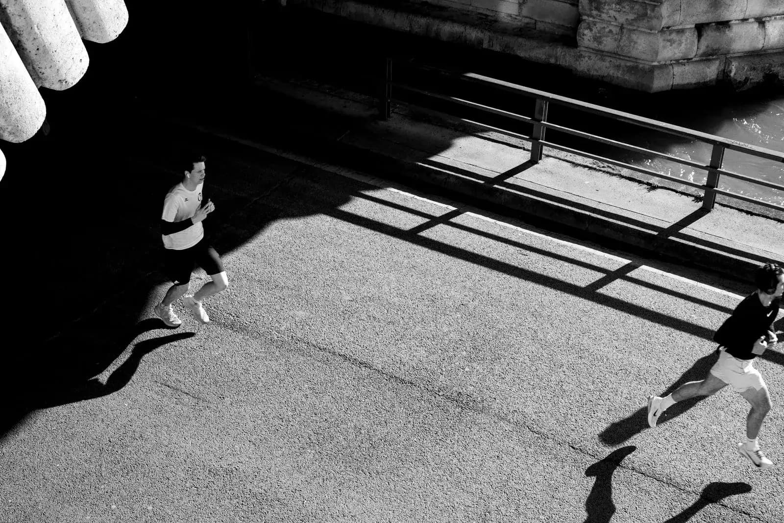 Two runners crossing a Paris riverside path beneath a bridge, in hard sunlight and shadow.
