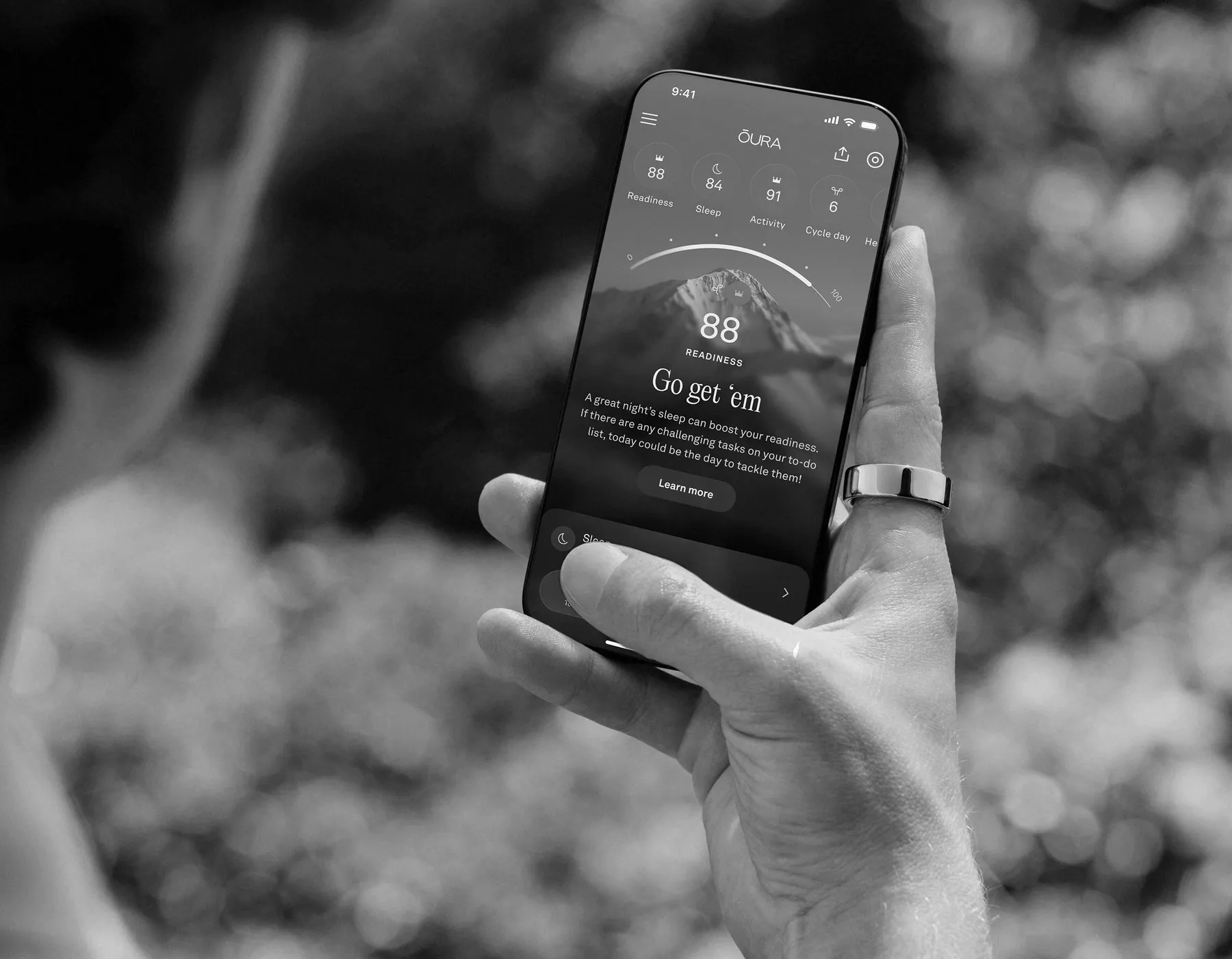 Hand wearing an Oura Ring holding a phone showing the Oura app readiness score